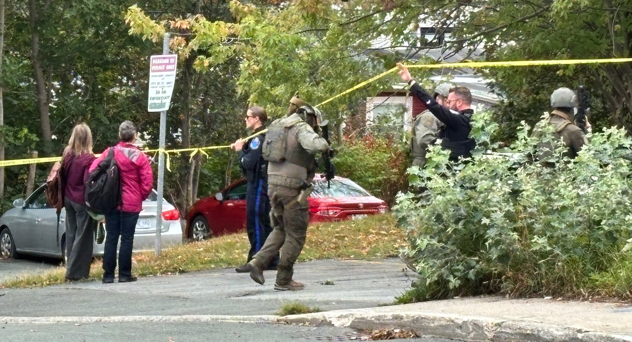 Police in tactical gear walk under police tape, while onlookers watch.