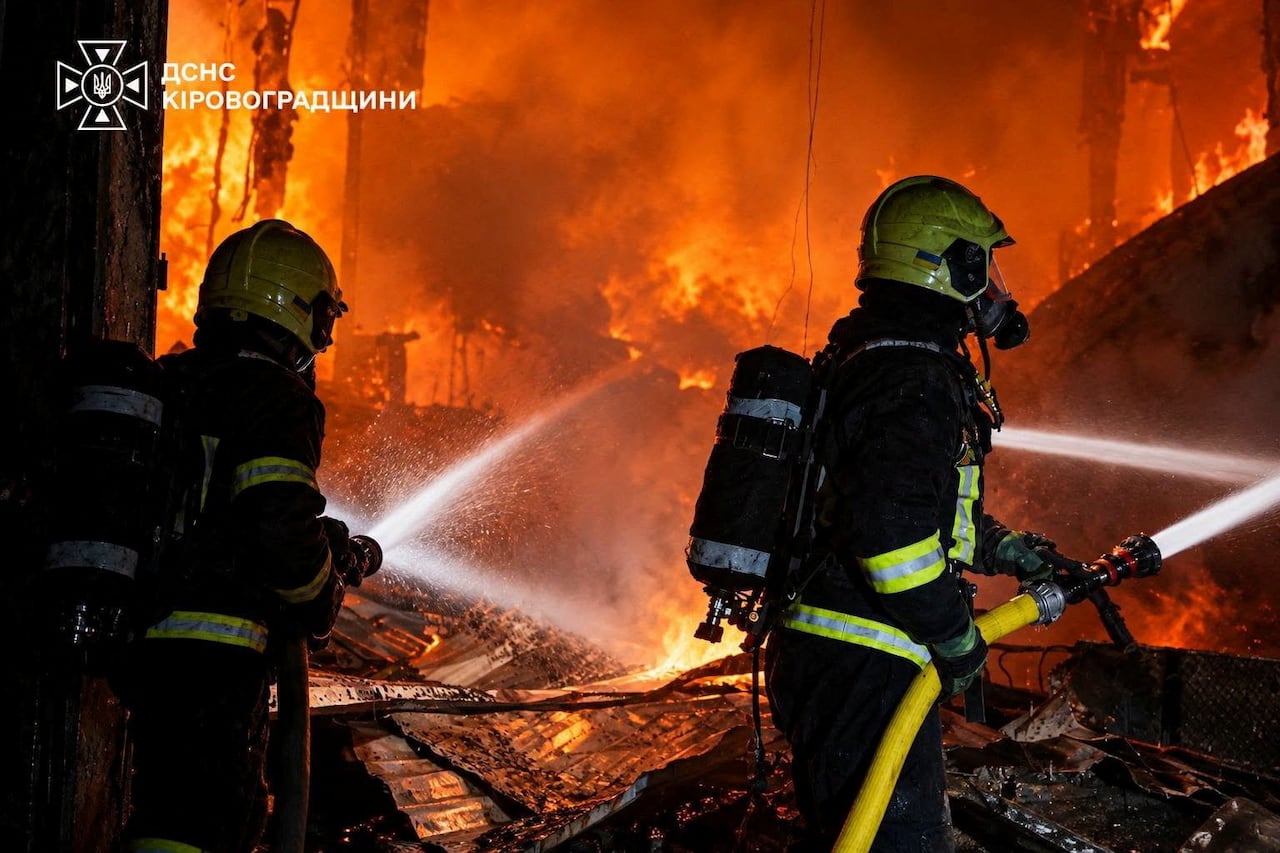 Two firefighters are shown in the foreground, with orange flames dominating the background of the picture.