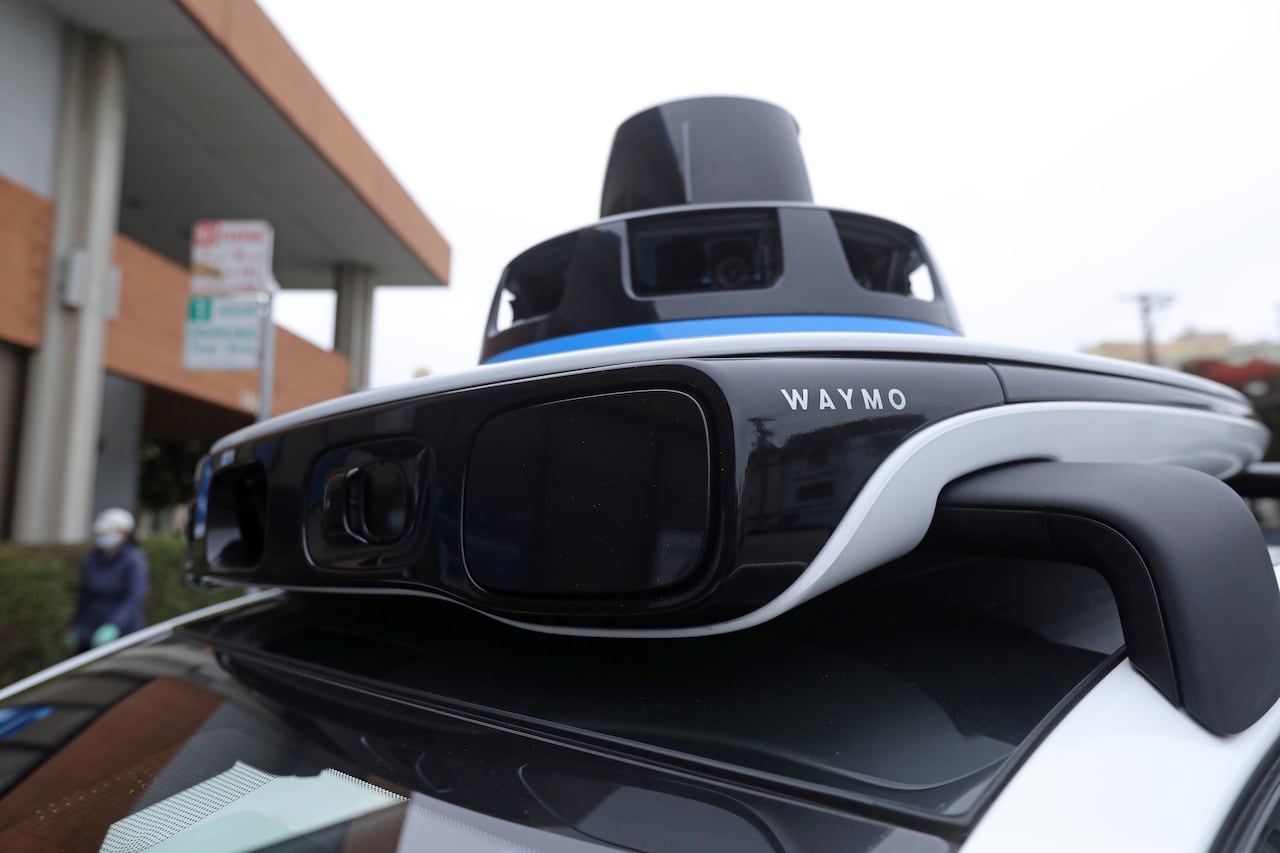 a piece of technology with a camera that says "waymo" sits atop a white car