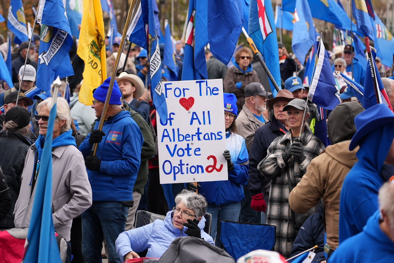 A group of people with Alberta flags stand together. A woman in the blue jacket holds a sign in the centre of the photo.