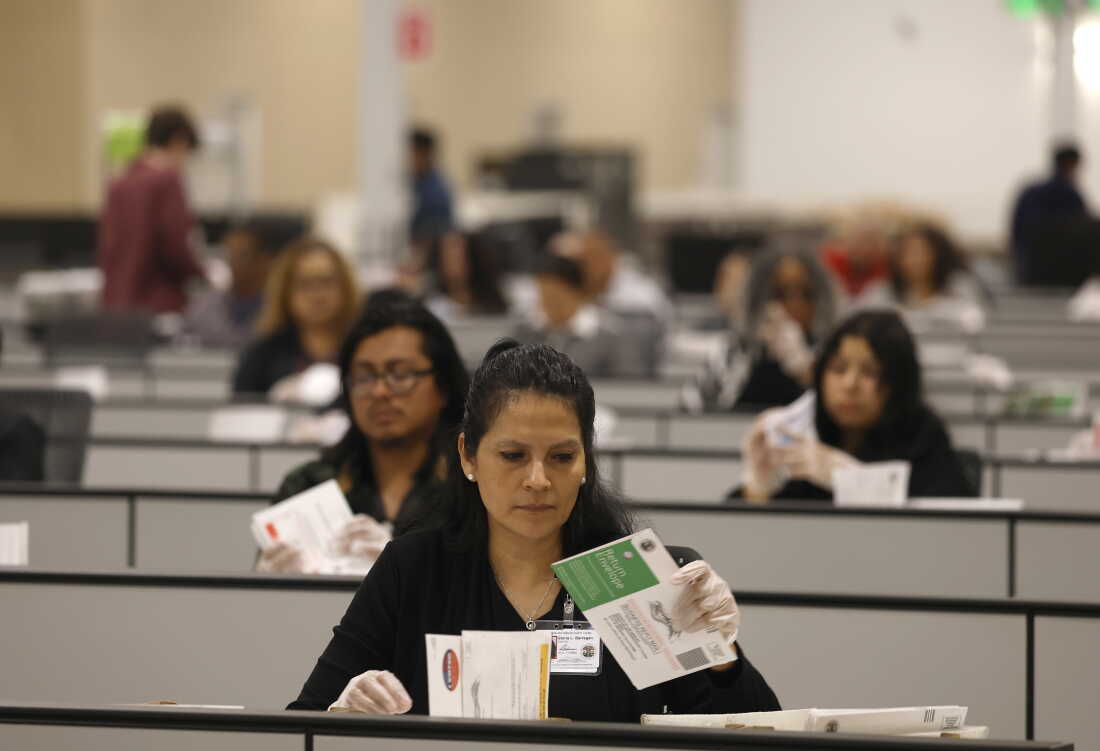 Election workers who are seated and wearing clear gloves process ballots for the California state primary.