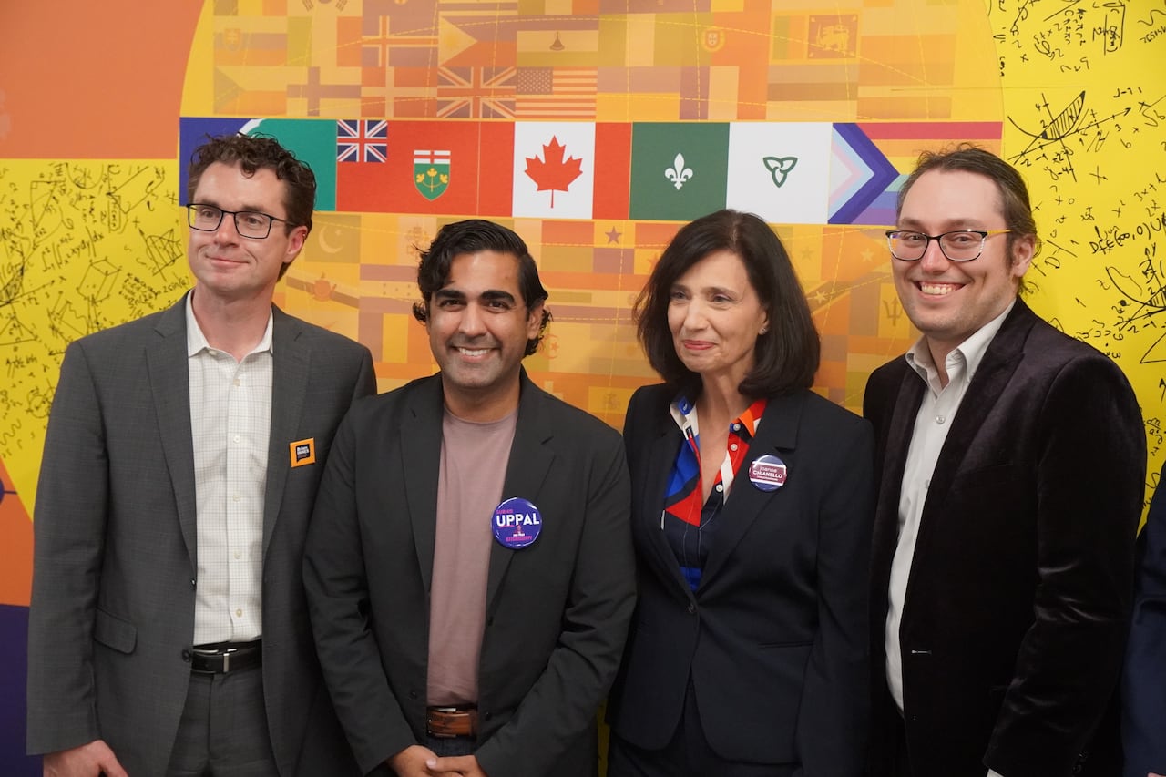 A group of four people stand smiling in front of an orange background.