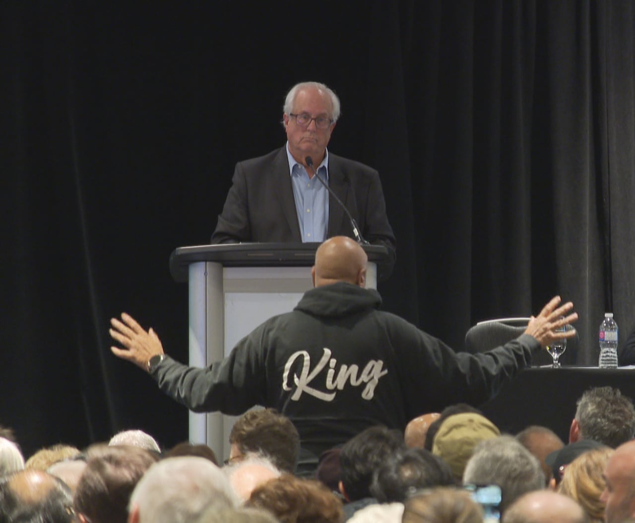 At a large public gathering, a man wearing a sweatshirt that says 'King' raises his arms while speaking to a man at a podium.