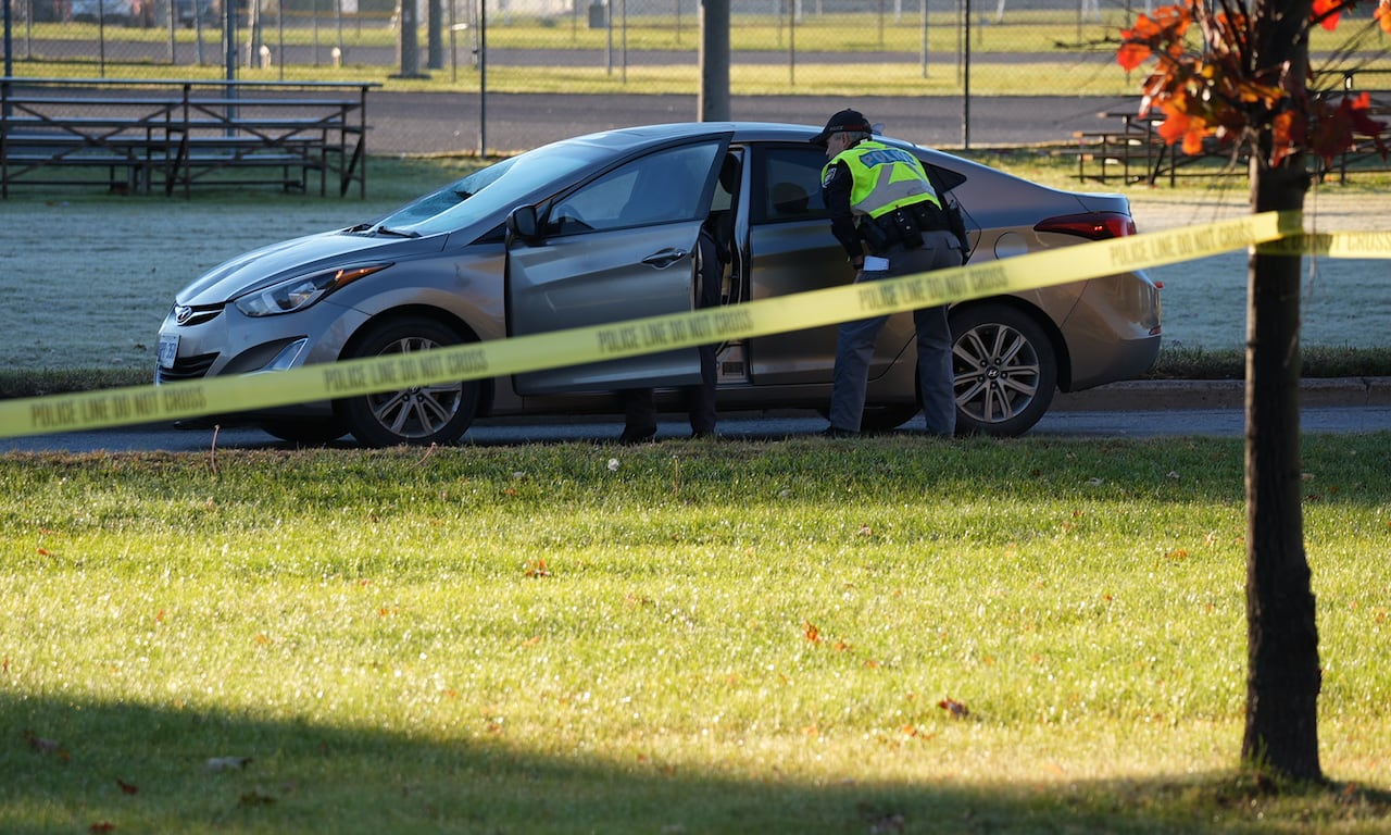 Crash investigators look at a stopped sedan on a closed road. It's a sunny autumn morning.