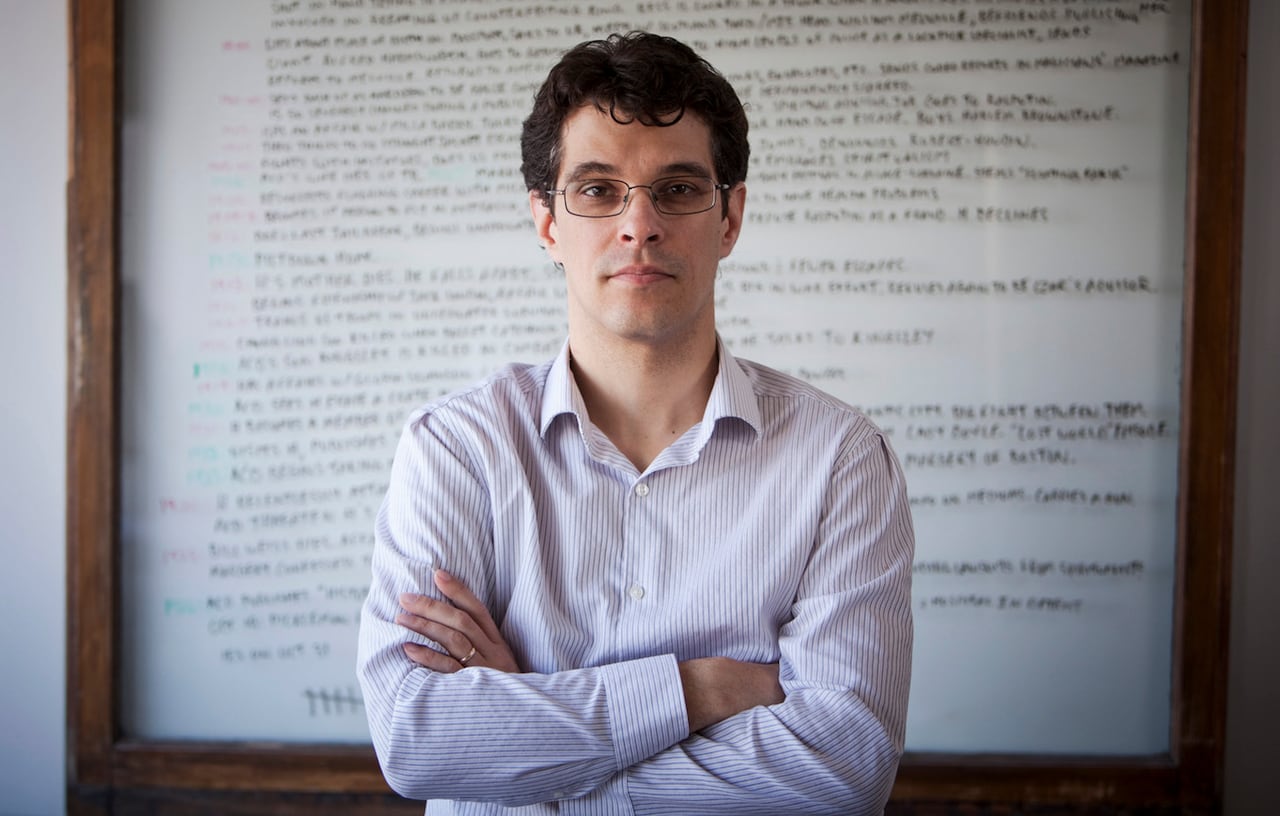 A man stands with his arms crossed looking at the camera in front of a whiteboard.