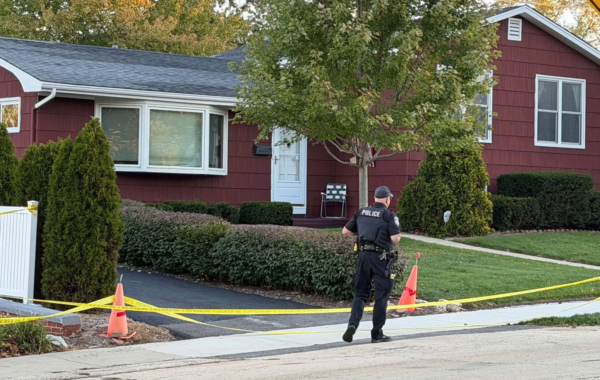 A Mundelein police officer walks past crime scene tape and traffic cones toward a red ranch home