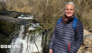 Eleanor stands by a waterfall. She has white hair, which is tied back, and wears a navy coat. She is smiling at the camera.