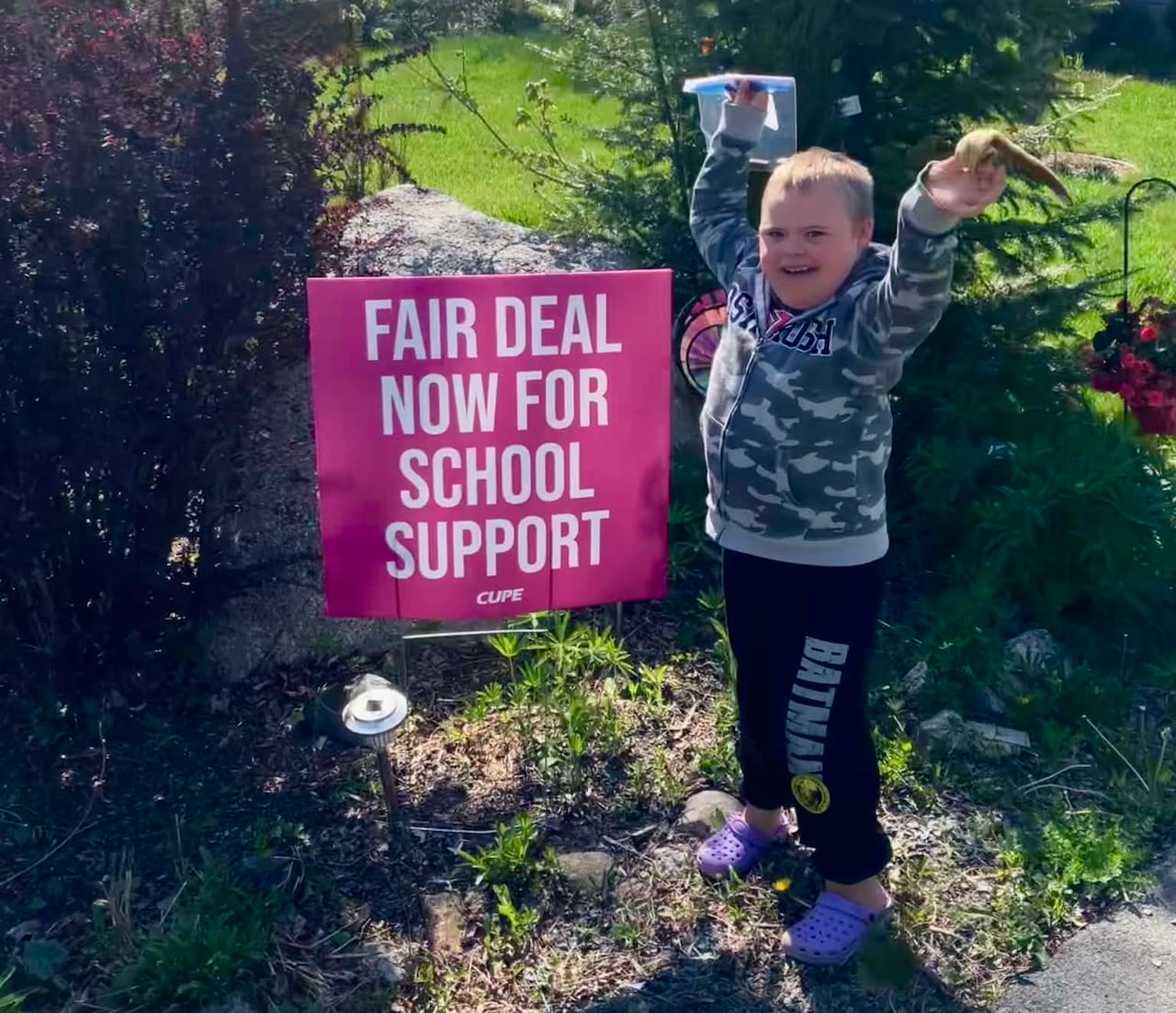 A young boy with down syndrome poses next to a pink yard sign that says "Fair deal now for school support."