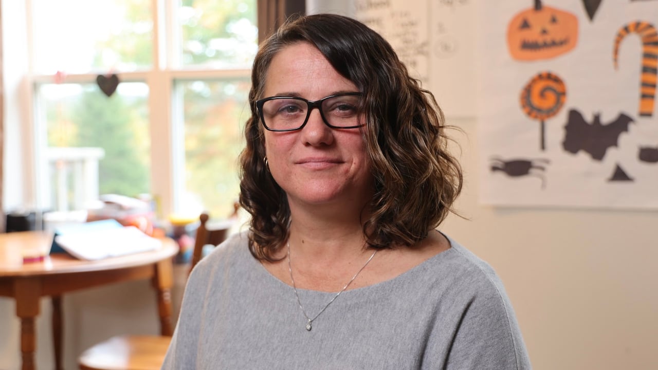 A white woman with short brown curly hair wears glasses and a grey shirt.