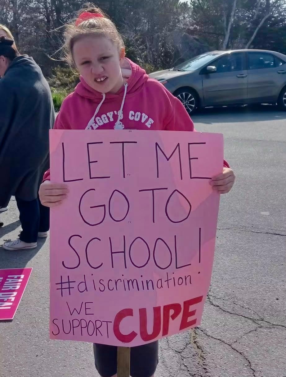A young white woman stands outside wearing all pink. She's holding a poster that says "let me go to school! #discrimination. we support CUPE."