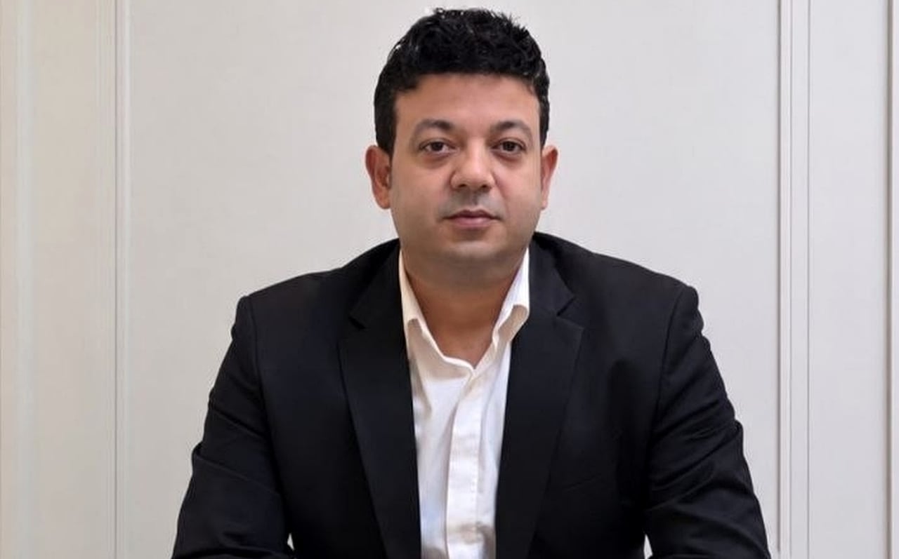 A man in a black suit looks forward with a serious expression.
