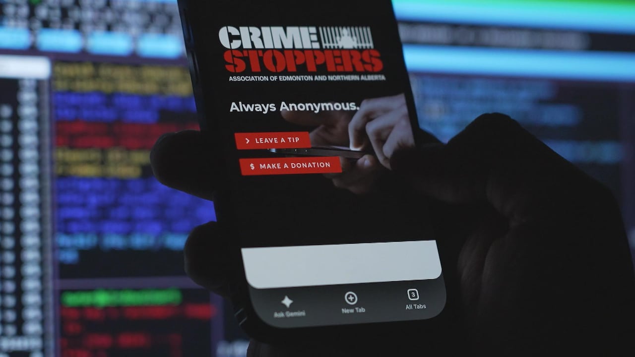 A phone displays the Crime Stoppers webpage, in front of a computer screen.