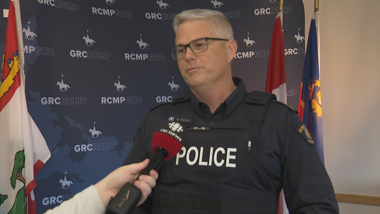 A man in glasses and a uniform with 'police' in white font on the front speaks into a black and red microphone.