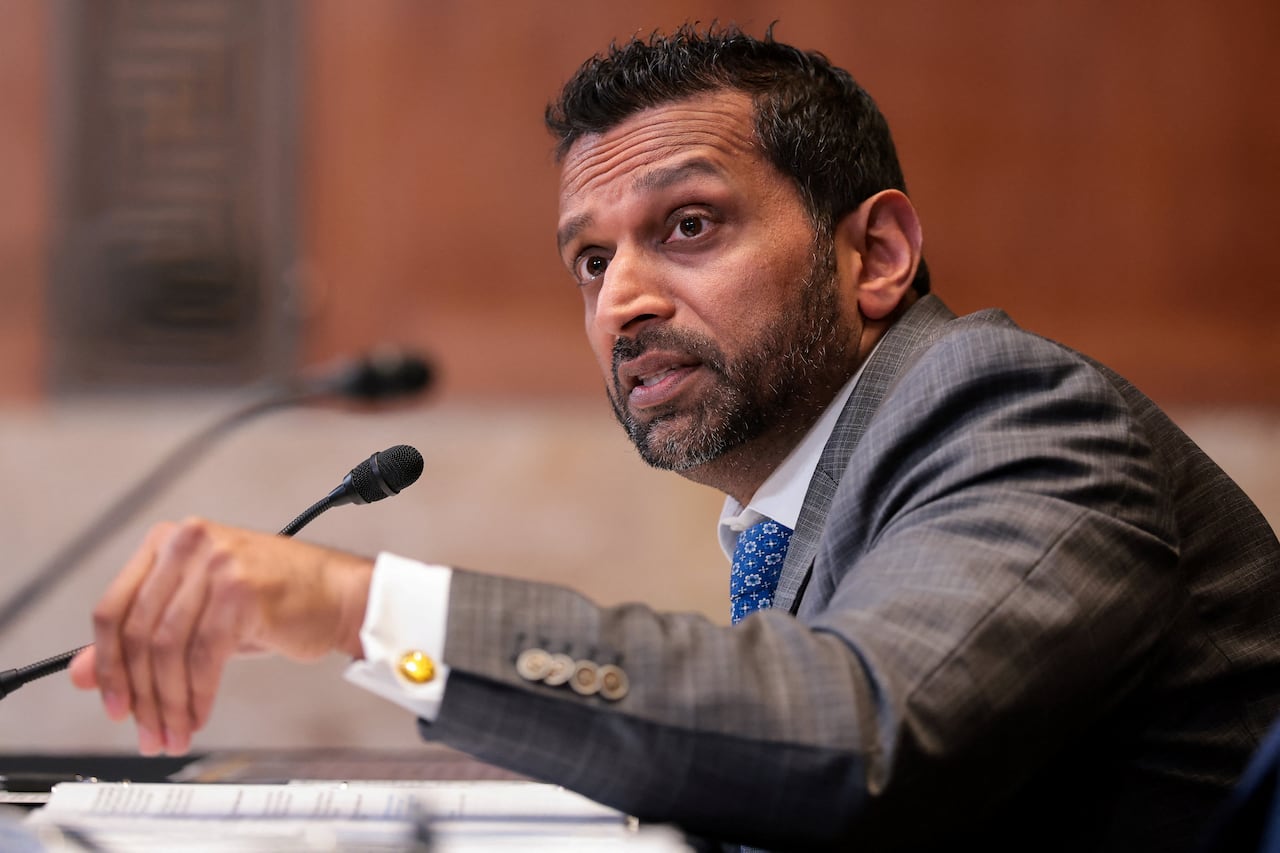 A man in a grey suit and a blue tie seated, speaking at a microphone