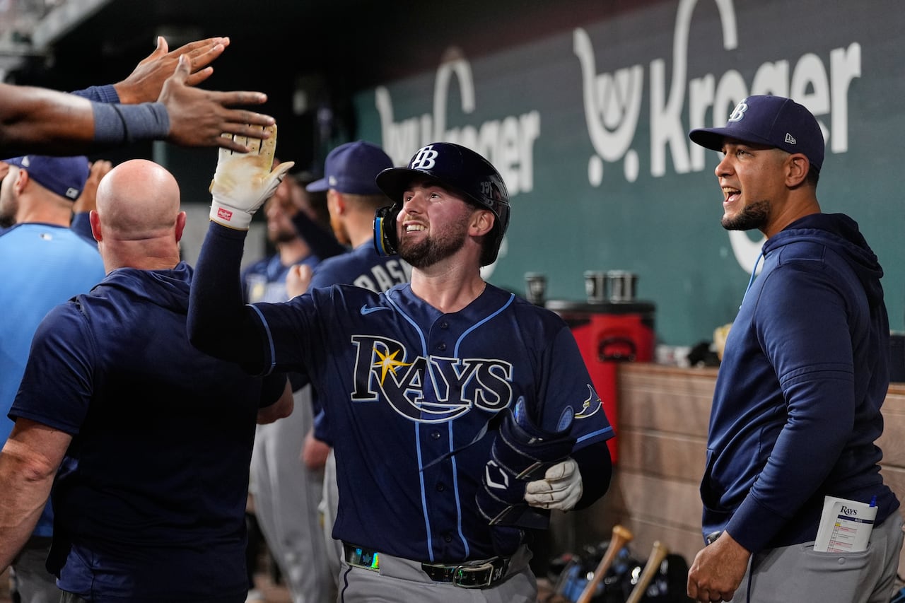 A man wearing a baseball helmet and a navy jersey spelling "Rays" high fives a teammate in the baseball dugout.