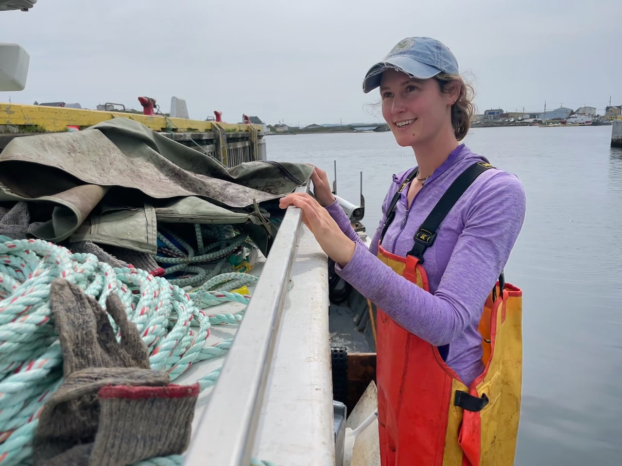 Lillian Saul, wearing a worn ball cap and bright yellow and orange oilskins, smiles while looking out over her fishing vessel