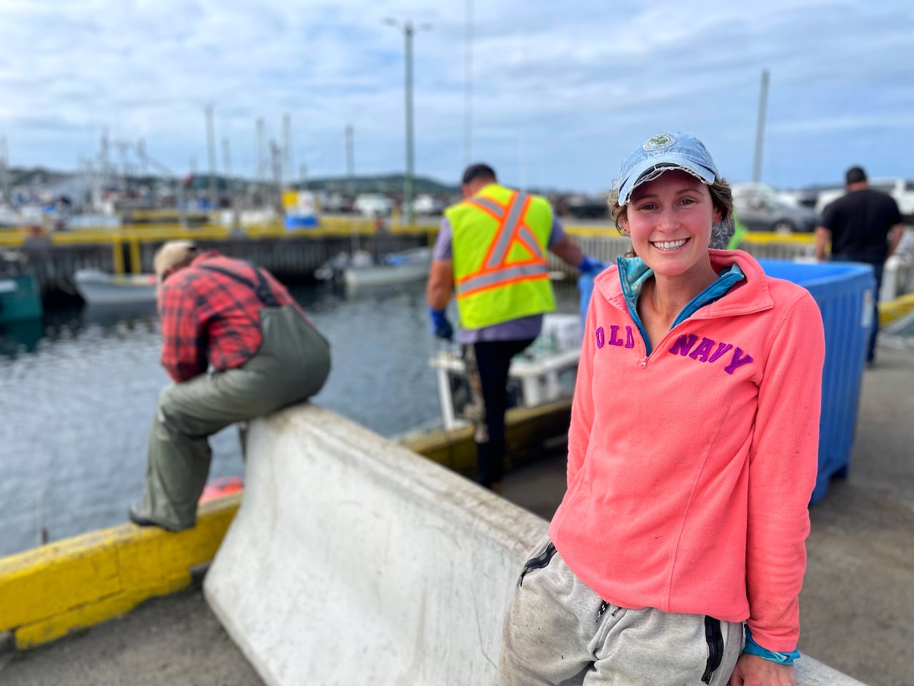 Lilly Saul leans on a concrete block, smiling into the camera. Behind her, men can be seen, wearing plaid, oilskins and safety vests.