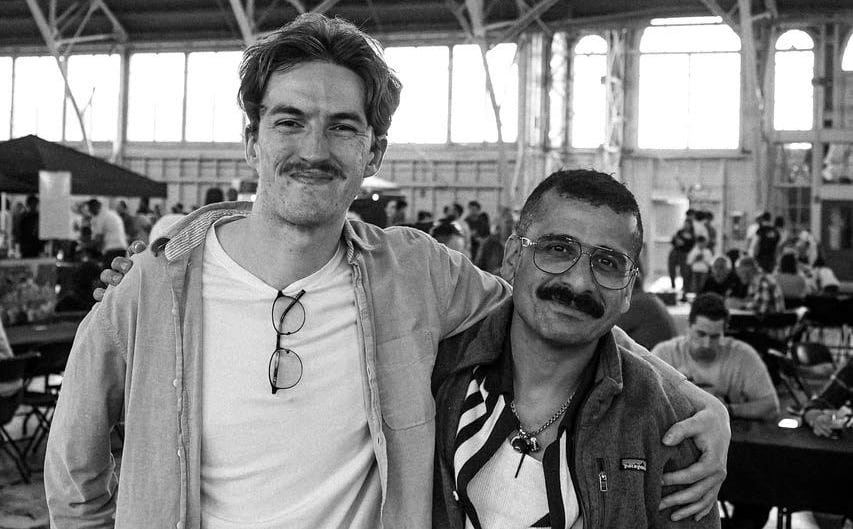 A tall man puts his arm around a shorter man. The pair smile and pose for a photo.