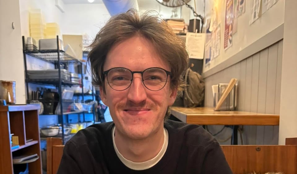 A white man with a moustache and glasses sits at a table, crosses his arms, and smiles.