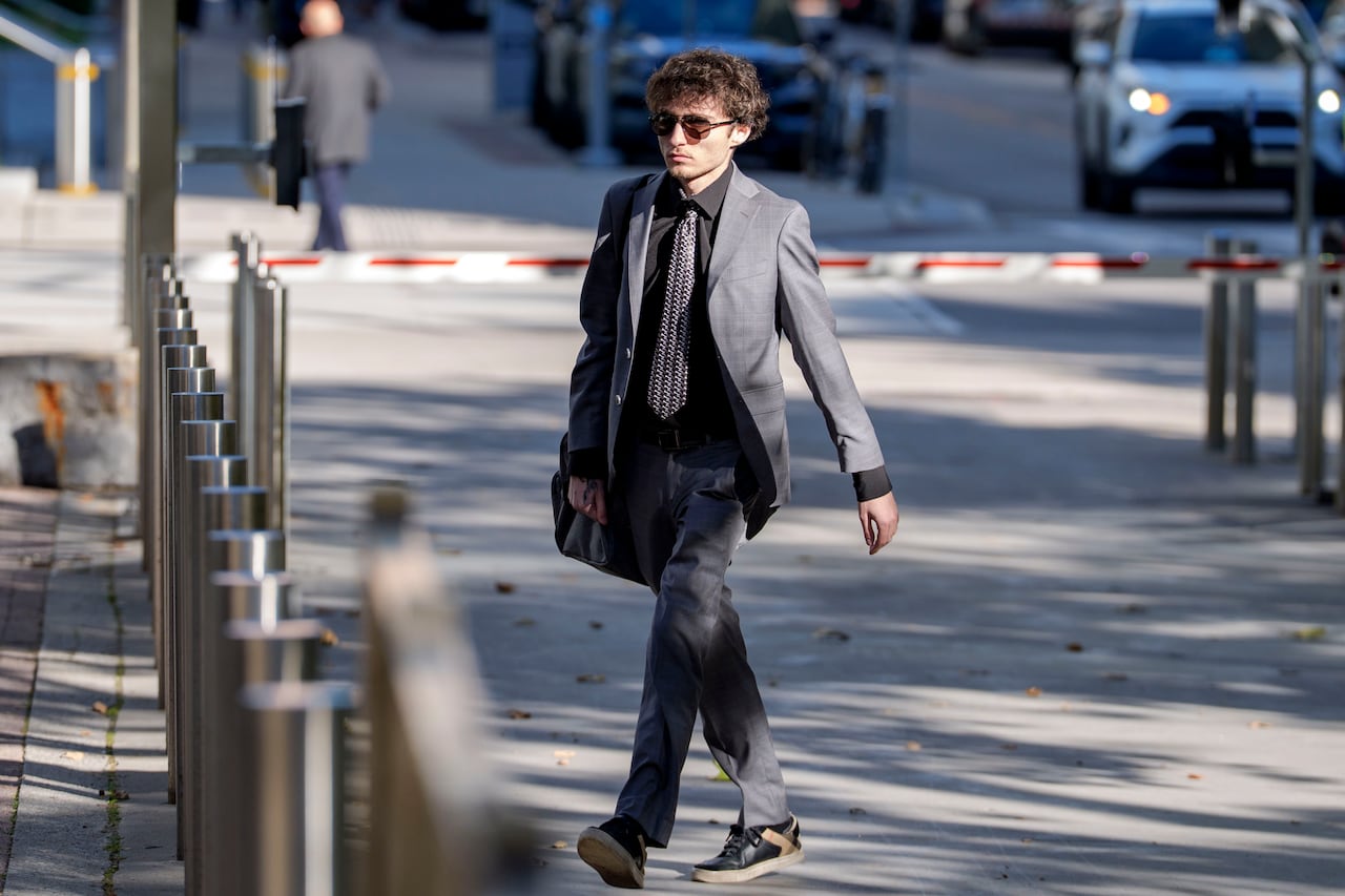 A man wearing a suit and sunglasses walks across a street in downtown Toronto outside a courthouse.