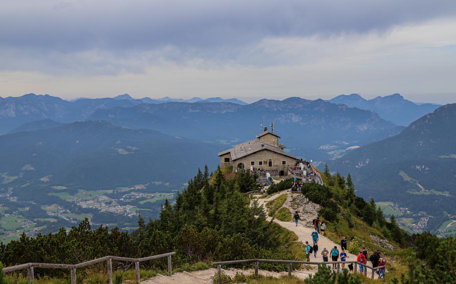 A stone building sits on a mountaintop as people walk up a gravel path through pine trees, with mountains in the distance. 