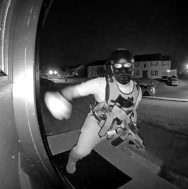 A doorbell camera view of a person wearing a mask and a tactical vest while carrying a rifle.