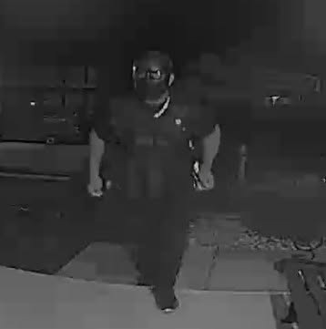 A grainy, low-light security camera image of a person wearing a mask and tactical vest approaching a home.