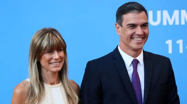 A medium shot shows Pedro Sanchez and his wife Begona Gomez standing together, while they are both smiling. Begona is wearing a white and cream coloured top and has long blonde hair with a fringe. Pedro is wearing a black suit, white shirt and blue tie. He has short black hair which is swept upwards/