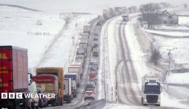 A road covered with snow and ice with snowy fields on either side, with a queue of cars and lorries in one direction and two lorries moving in the other.