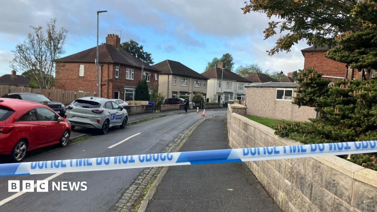 A police cordon blocks access to a residential street lined with semi-detached houses. Blue-and-white police tape stretches across the road in the foreground, while traffic cones and a person can be seen further down the street.