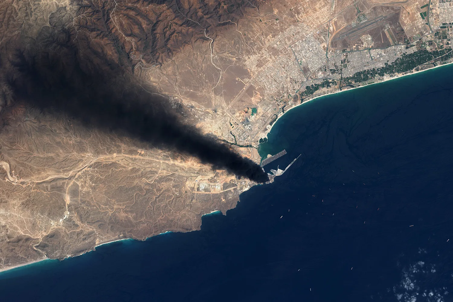 Aerial satellite footage shows a coastline where a thick plume of black smoke rises from a port area and stretches inland over mountainous terrain.