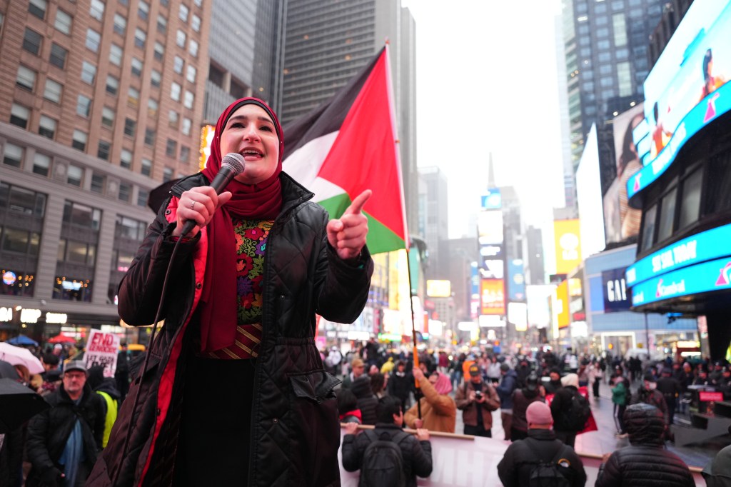 Linda Sarsour speaks into a microphone at a rally in Times Square in front of a Palestinian flag.