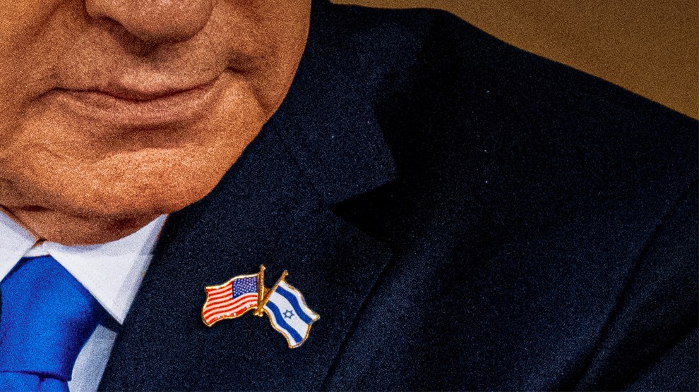A close-up of a man, seen from nose down to mid-chest, wearing a suit and blue tie. He wears a lapel pin with the American and Israeli flags. His left hand is caught mid-gesticulation, as though he’s just made a point.