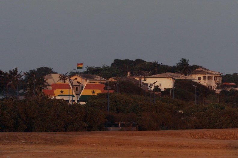 A Ghanian flag waves on a pole over homes where two images of the flag is seen down below.