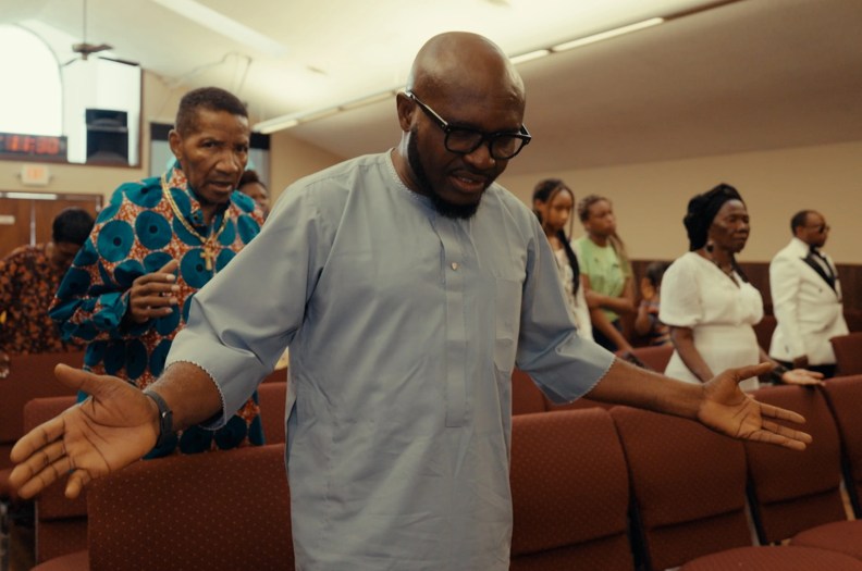 A man in the foreground opens his arms as he prays while several people stand in the rows behind him.