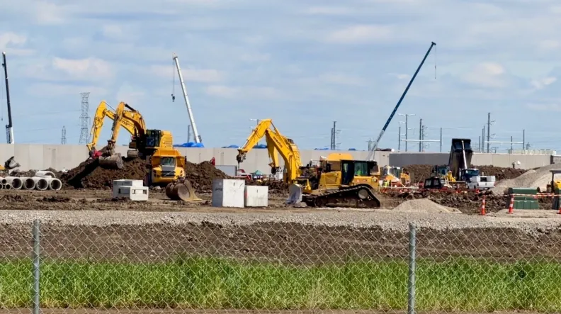 Four yellow construction excavators, three cranes and a dump truck sit in a lot behind a chainlink fence.