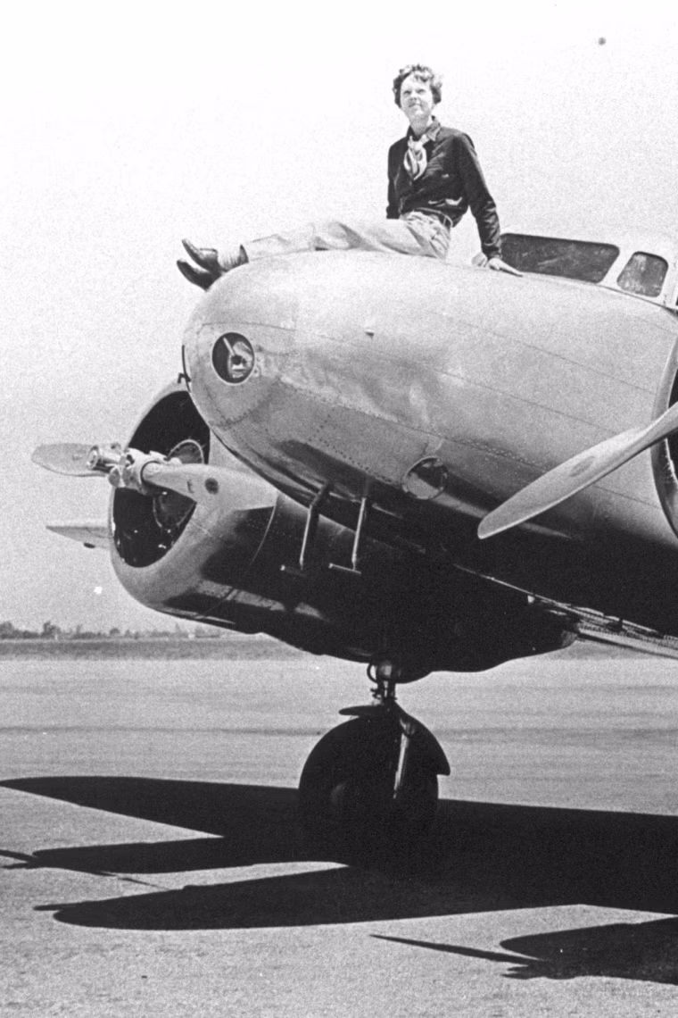 A black and white photograph of Amelia Earhart seated on the nose of a propeller plane.