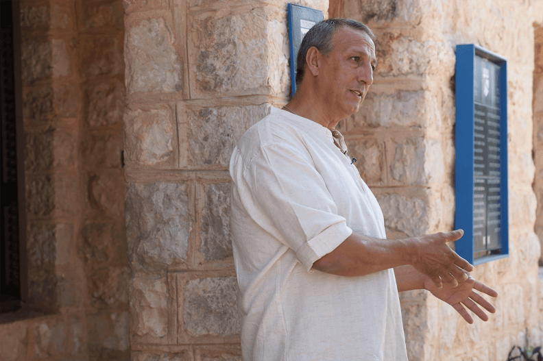 A man in a white shirt seen from the waist up gestures with both hands while speaking. He stands in front of a stone building.