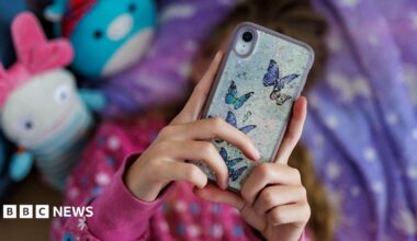 A young girl with long brown hair lying on her bed with a purple and white duvet, looking at her phone over her face, which has butterfly stickers on the back. There are two out-of-focus toys next to her in blue and white, and she is wearing a pink jumper with white and blue flowers on it.