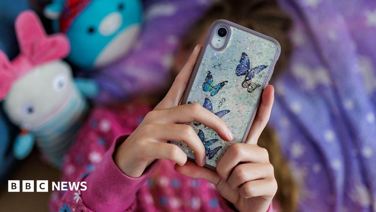 A young girl with long brown hair lying on her bed with a purple and white duvet, looking at her phone over her face, which has butterfly stickers on the back. There are two out-of-focus toys next to her in blue and white, and she is wearing a pink jumper with white and blue flowers on it.