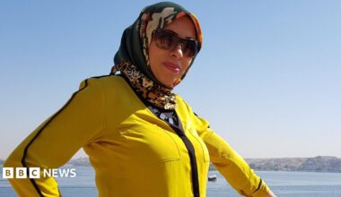 A woman is wearing a yellow jacket and a multicoloured headscarf. She is wearing sunglasses and standing in front of the ocean.