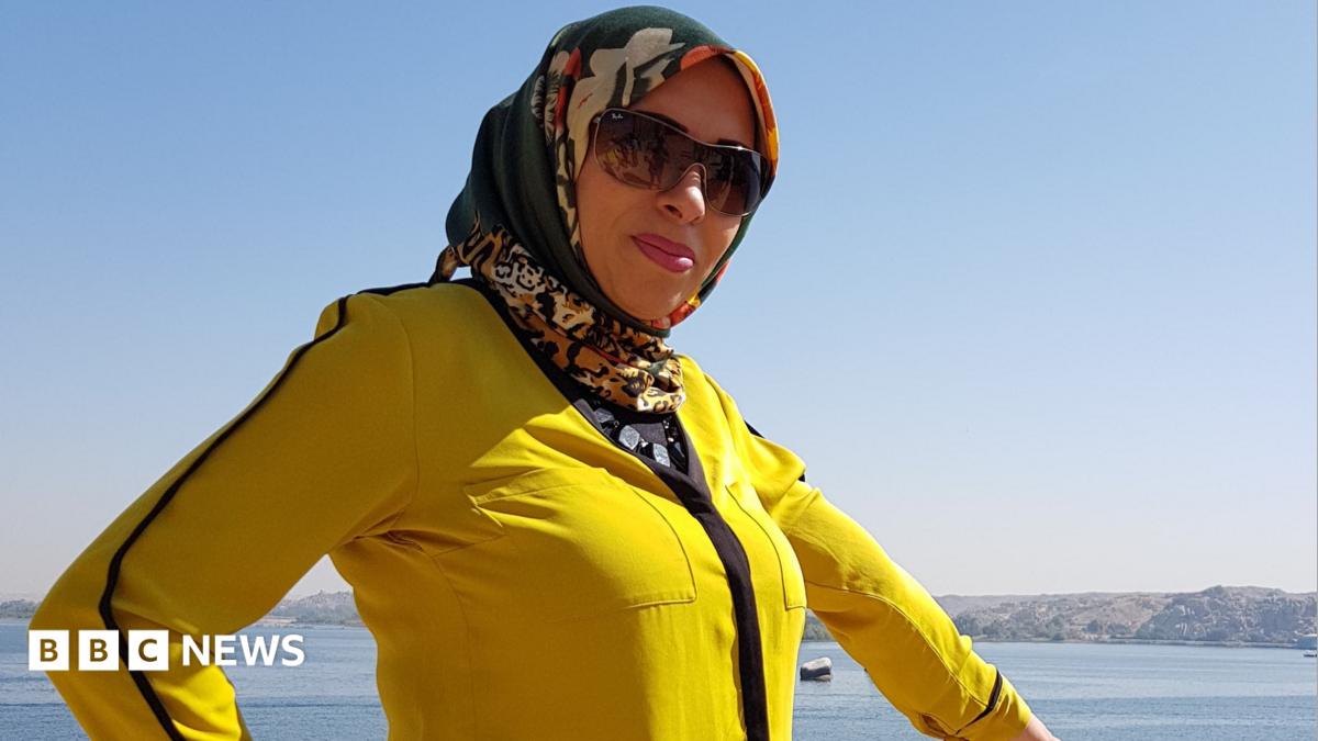 A woman is wearing a yellow jacket and a multicoloured headscarf. She is wearing sunglasses and standing in front of the ocean.