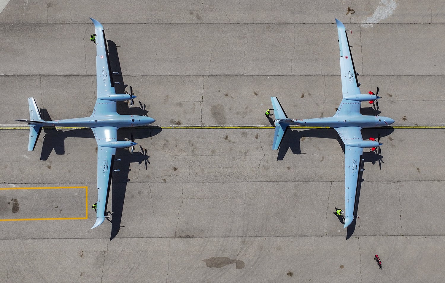 A top-down aerial view shows two large grey uncrewed aircraft with long wings parked one right in front of the other on an asphalt tarmac.