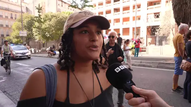 A medium shot shows Joanna talking into a BBC branded microphone while walking in the street during a protest in Marseille