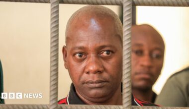 A close shot of a bald man looking through the bars of a cell