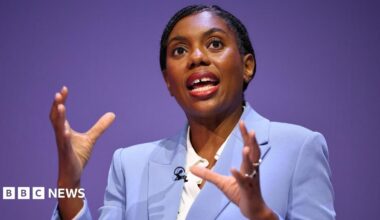 Kemi Badenoch speaking while wearing a light blue suit and white shirt. She's gesticulating with her hands as she speaks.