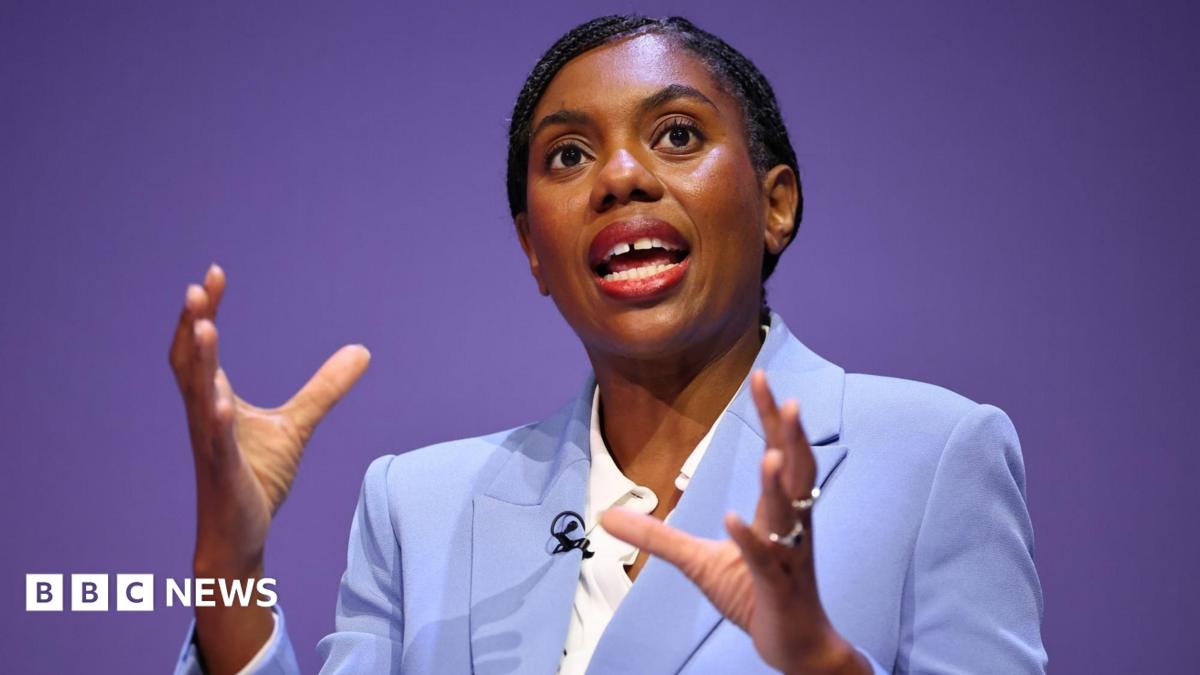 Kemi Badenoch speaking while wearing a light blue suit and white shirt. She's gesticulating with her hands as she speaks.