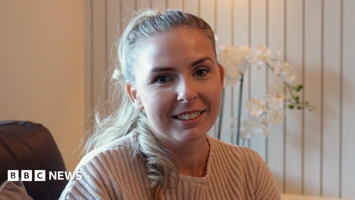 Kelly O'Shea sat in her living room, against a backdrop of a cream wall, with an orchid on the shelf behind her. She is wearing a cream ribbed top and has blonde hair tied back from her face.