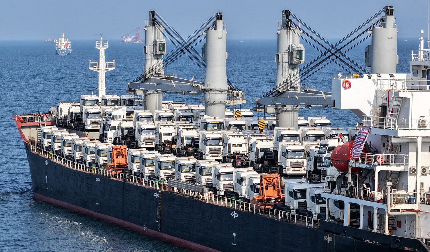 A large cargo ship traveling across open water, its deck fully loaded with rows of white semi-truck cabs.