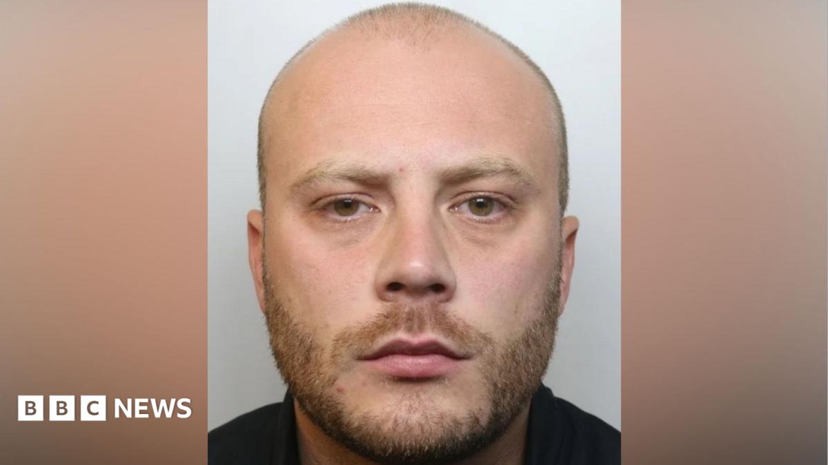 A police mugshot of a balding man in his late 20s, with light stubble and brown eyes, looking into the camera with a serious expression.